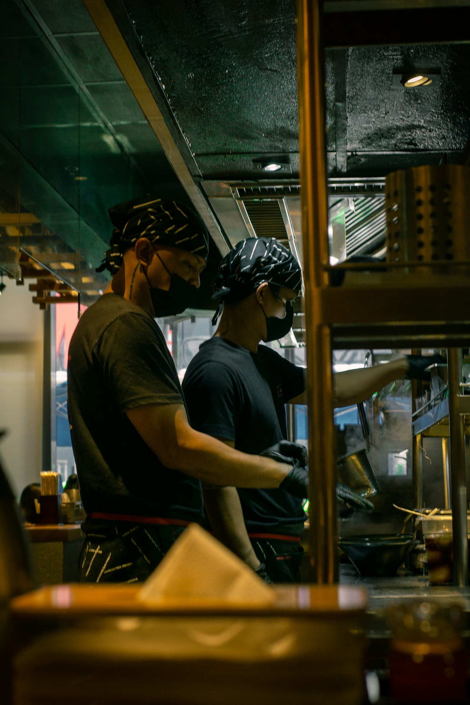 두건을 쓴 두 요리사가 좁은 라면 가게 주방에서 나란히 서서 조리하고 있다