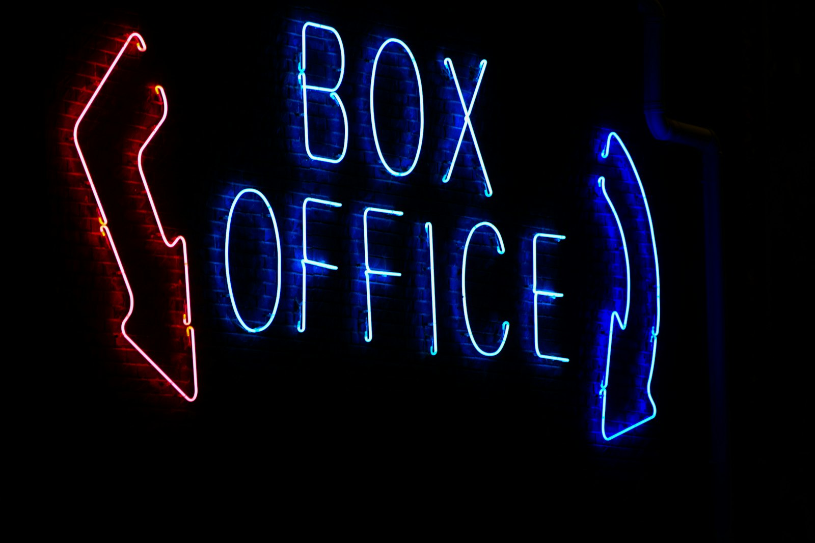 어두운 벽돌 벽에 붉은색과 파란색 네온으로 'BOX OFFICE'와 양쪽 화살표가 켜져 있다