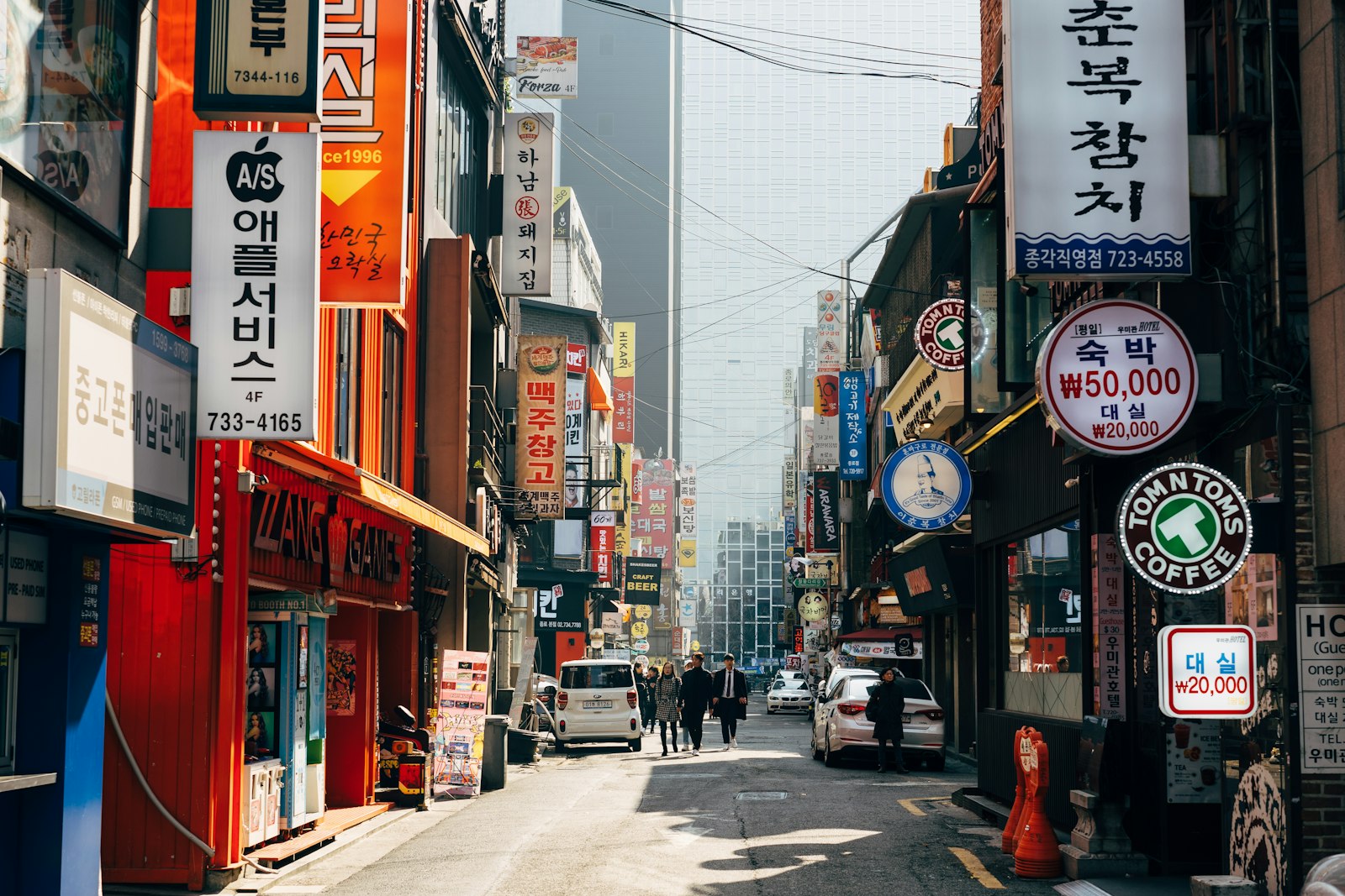 겨울 햇살이 비스듬히 든 서울 도심 골목, 한글 간판과 '숙박 ₩50,000' 표시가 빼곡한 거리를 몇 사람이 걸어간다